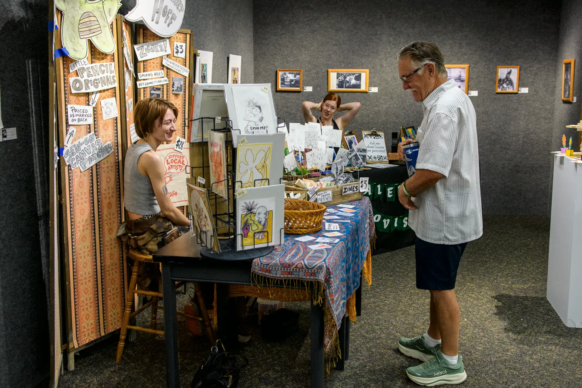 Artists at a table of prints and zines talking with a visitor indoors
