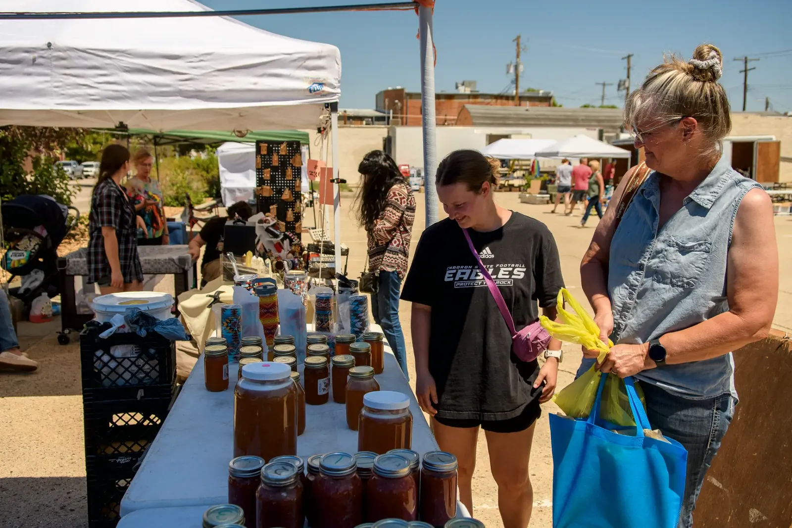 Shoppers at a table of canned jars and tumblers under market canopies